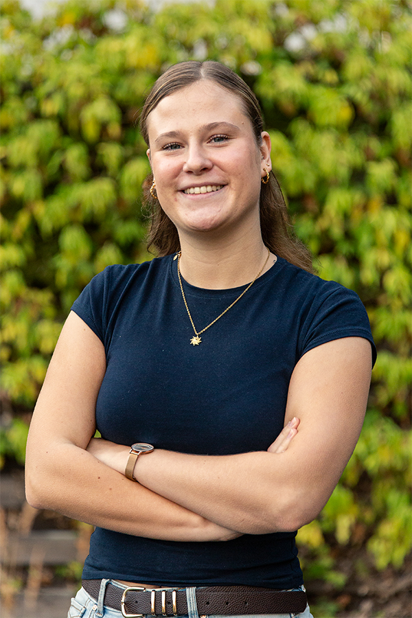 Junge Frau in blauem T-Shirt lächelt mit verschränkten Armen vor grüner Hecke, WWWEWERS Marketingagentur.